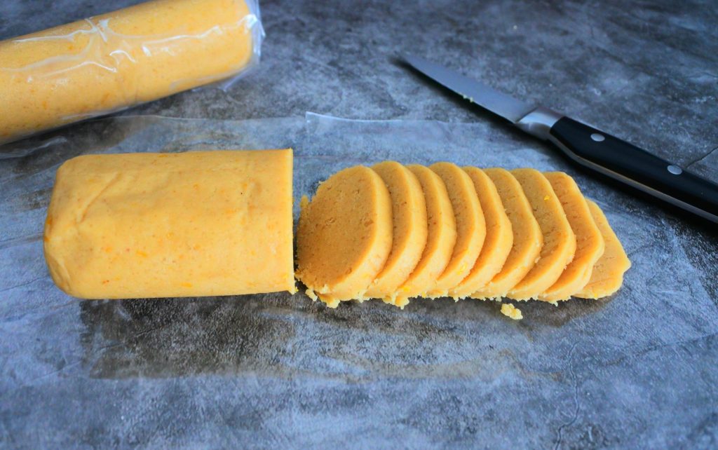 An angled image of a log of chilled lemon shortbread cookie being sliced into rounds before being baked