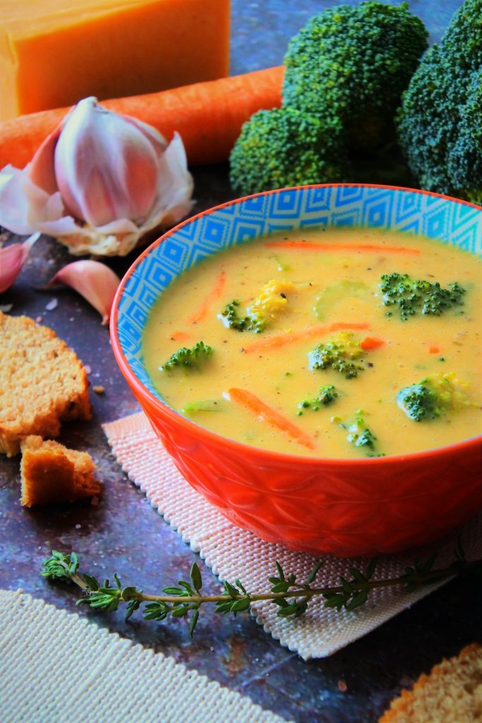 An angled head on image of a bowl of broccoli cheddar soup surrounded by fresh broccoli, carrots, cheese, bread and herbs.