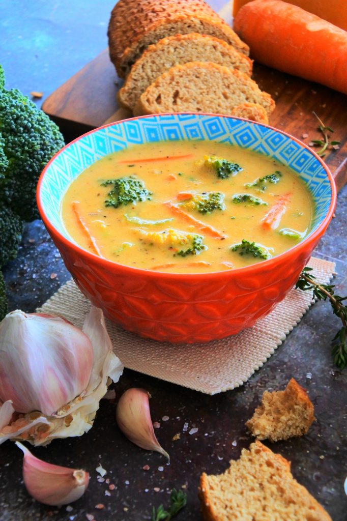 An angled head on image of a bowl of broccoli cheddar soup surrounded by fresh broccoli, carrots, bread and herbs.