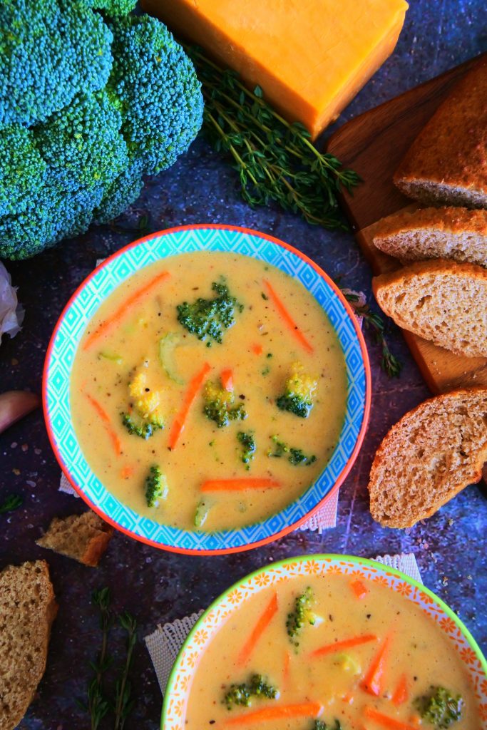 An overhead image of two bowls of broccoli cheddar soup surrounded by fresh broccoli, cheese, sliced bread and herbs.
