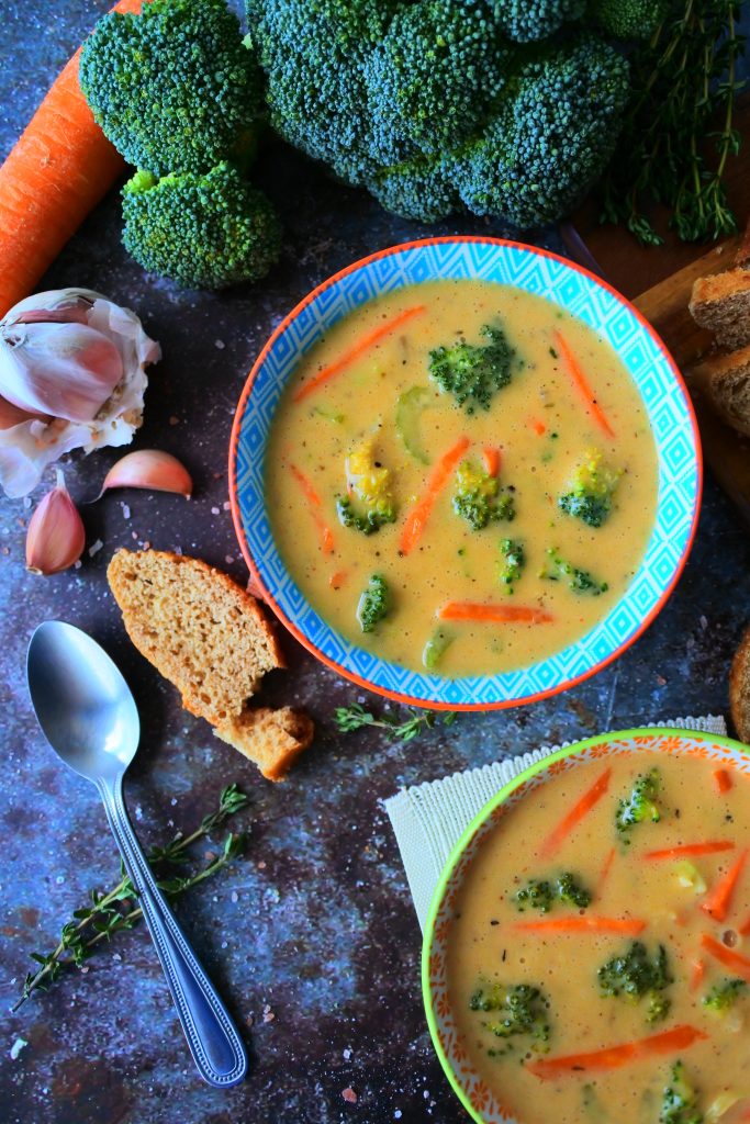 An overhead image of a bowl of broccoli cheddar soup surrounded by fresh broccoli, carrots, bread and herbs.