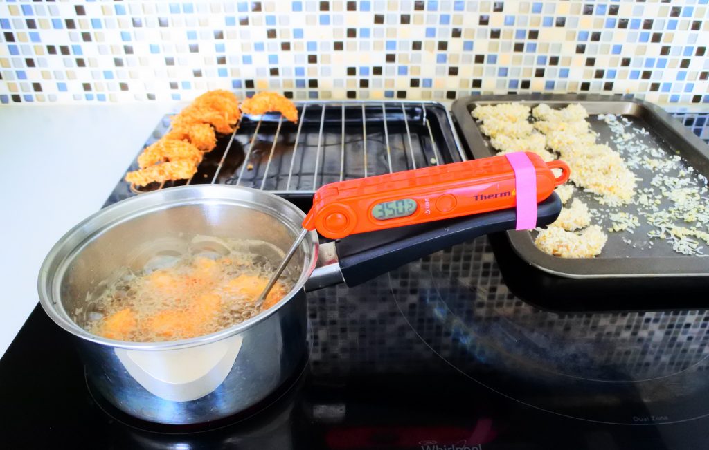 A head on image of a small pot with oil being and a batch of coconut shrimp being fried with a two trays in the background, one with fried shrimp and the other with unfried shrimp