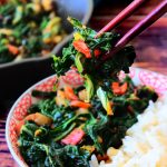 A head on image of swiss chard ohitashi between a pair of chopsticks with a bowl of ohitashi and rice in the background