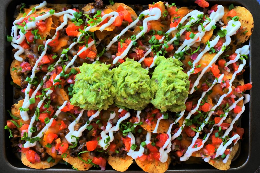 An overhead image of a tray cheesy beef nachos topped with salsa, sour cream, and guacamole