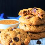 A head on image of a stack of peanut butter chocolate chip oatmeal cookies on a surface with the top cookie bitten into and a scattering of chocolate chips and raw oats around it plus a small dish of peanut butter behind it