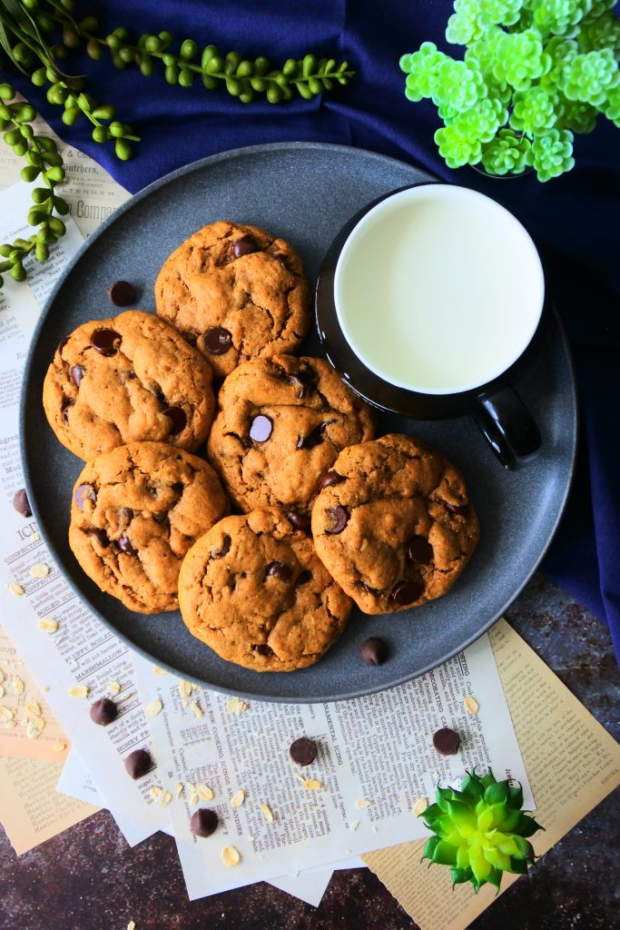 An overhead image of a plate of peanut butter oatmeal chocolate chip cookies and a mug of fresh milk surrounded by papers of recipes and various plants as well as oatmeal and chocolate chips and a dark blue linen cloth
