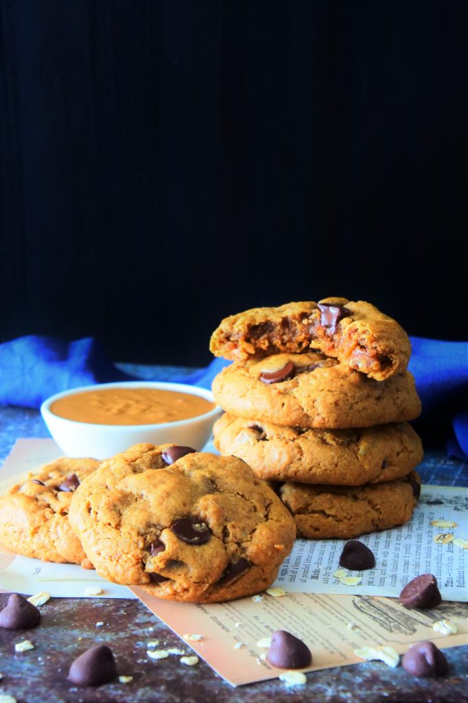 A head on image of a stack of peanut butter chocolate chip oatmeal cookies on a surface with the top cookie bitten into and a scattering of chocolate chips and raw oats around it plus a small dish of peanut butter behind it