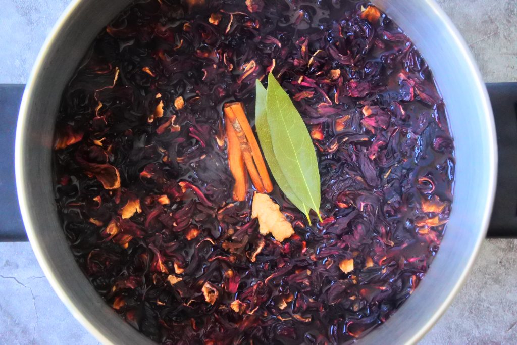 An overhead image of a pot of boiling water with dried sorrel/roselle added to it along with various herbs and spices including bay leaves, cinnamon sticks cloves and ginger