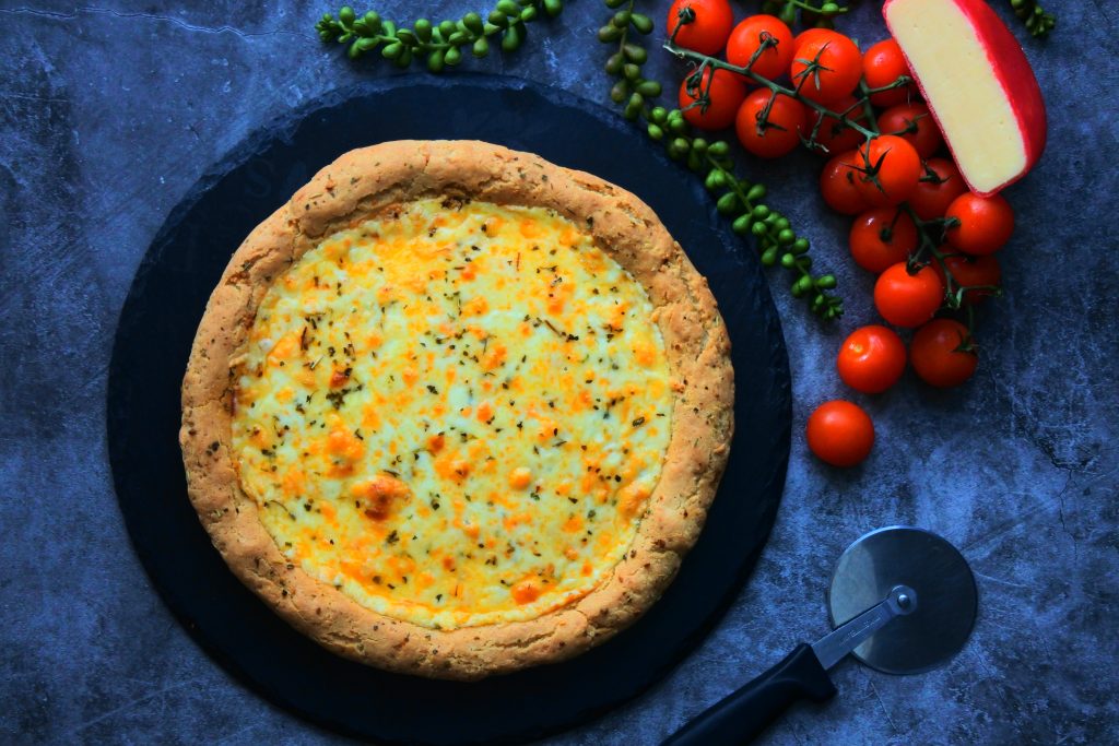 An overhead image of an unsliced freshly baked cheesy pizza on a slate serving plate with a pizza cutter next to it