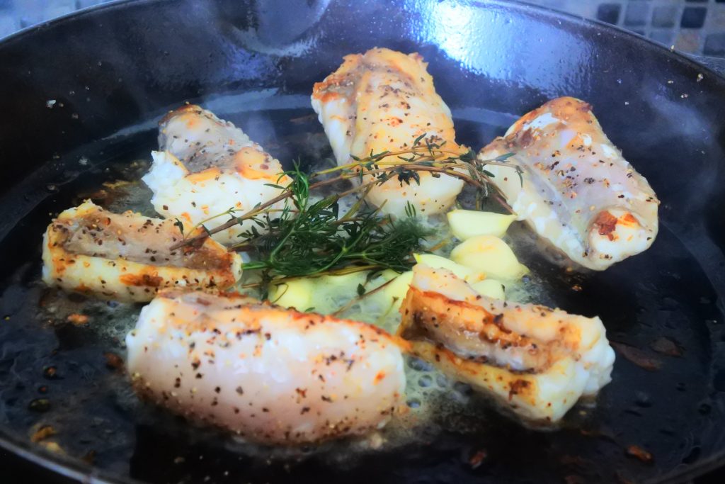 An angled image of monkfish fillets being pan fried in a skillet with garlic, butter and herbs in the middle
