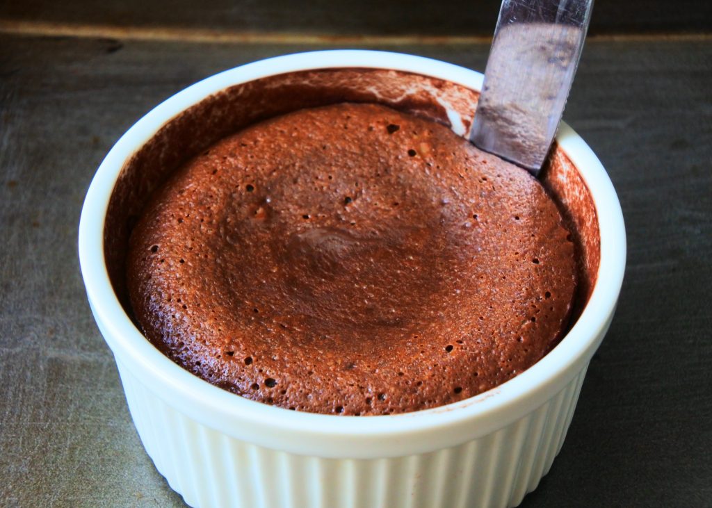 An angled close up image of a knife being run along the edge of a fresh baked molten chocolate cake while it's in the ramekin