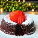 A close up image of a split molten chocolate cake topped with icing sugar and a fresh strawberry with greens and lights in the background