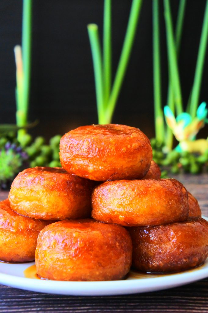 A close up image of a plate of stacked fried potato mochi after being brushed with sauce on a plate with greenery and a figurine in the background.