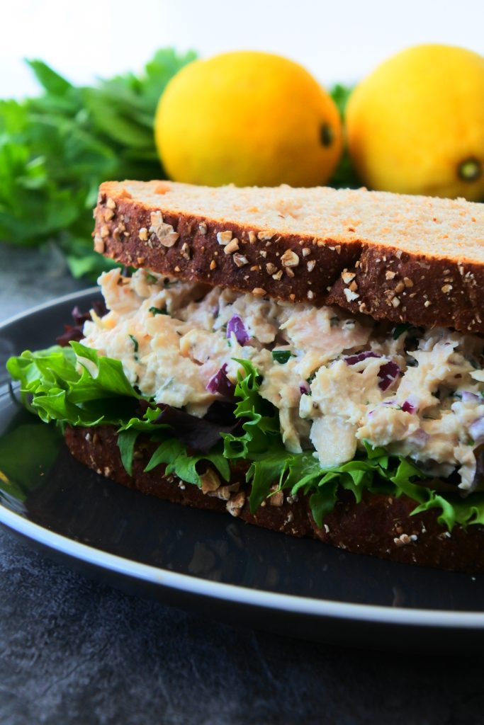 A close up image of a tuna salad sandwich on a plate with fresh produce in the background