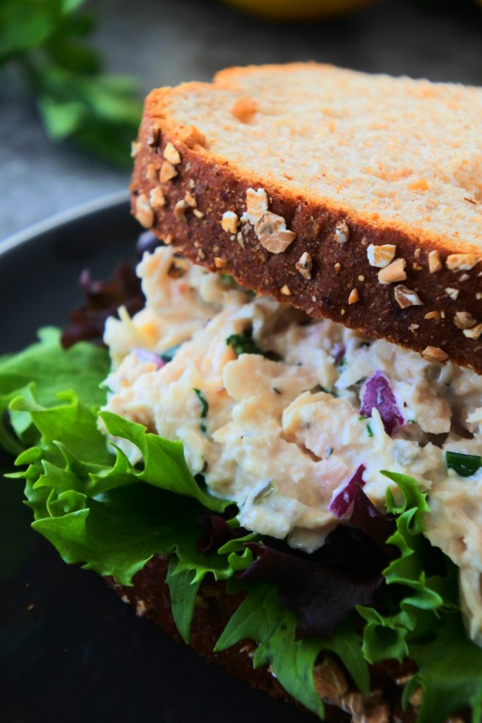 A close up image of a tuna salad sandwich on a plate with fresh produce in the background