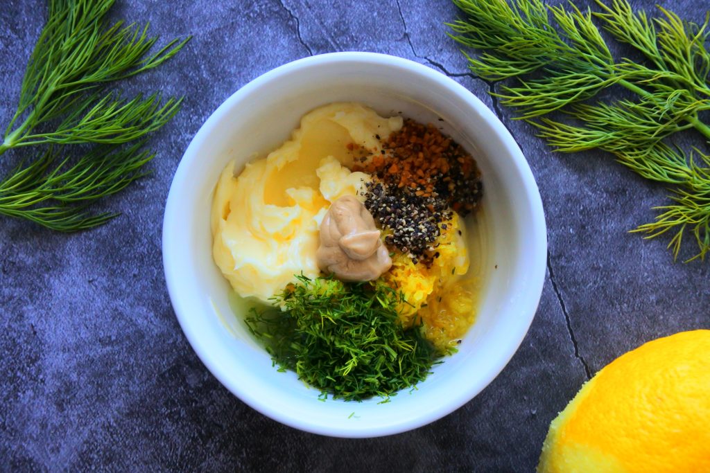 An overhead image of an unmixed batch of lemon dill butter in a white bowl surrounded by fresh dill and lemon