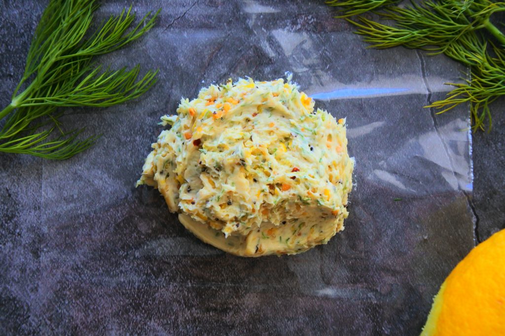 An overhead image of a mixed lemon dill butter scraped onto a piece of food grade plastic surrounded by fresh dill and lemon