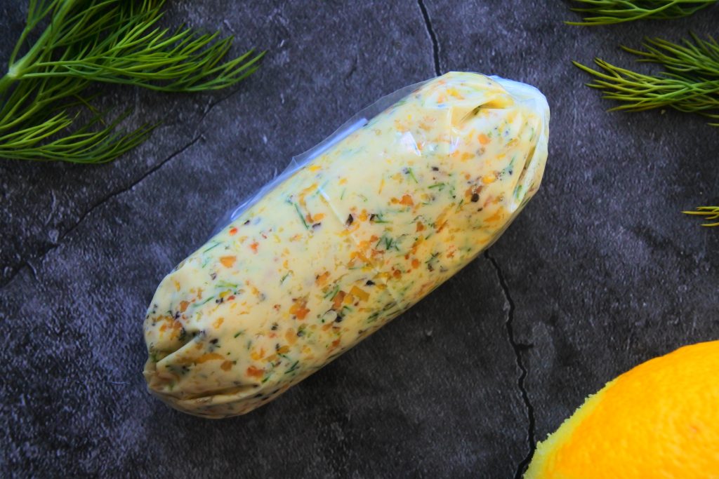 An overhead image of a log of lemon dill butter rolled in plastic surrounded by fresh dill and lemon
