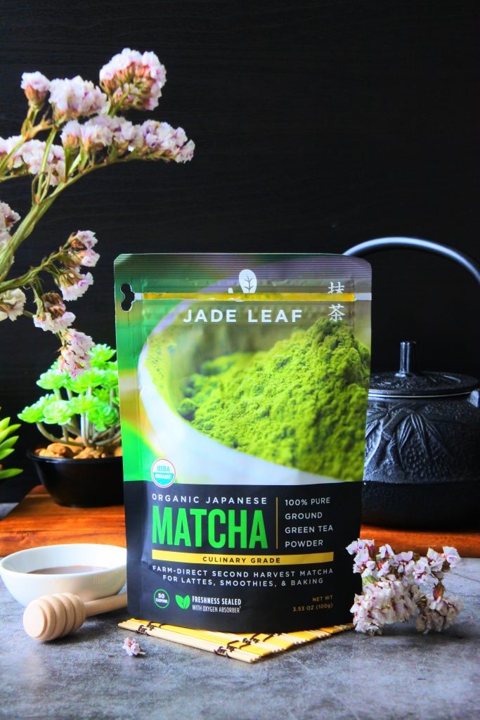 A head on image of a pack of Japanese culinary matcha surrounded by flowers, honey and a black tea kettle in the background