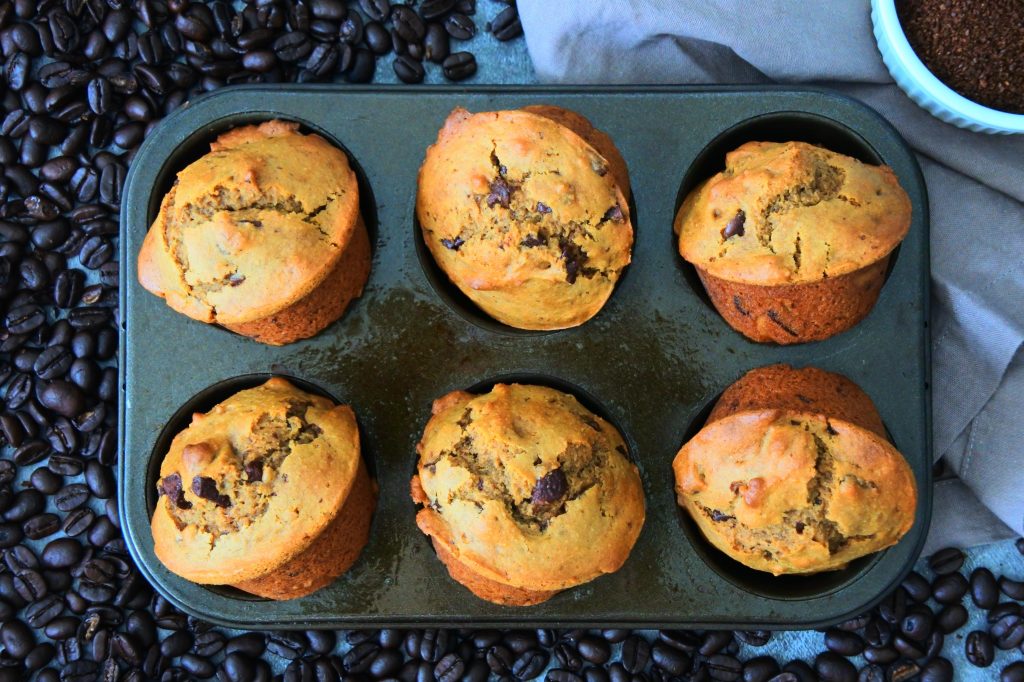 An overhead image of a freshly baked half dozen of chocolate chip coffee muffins surrounded by fresh coffee beans