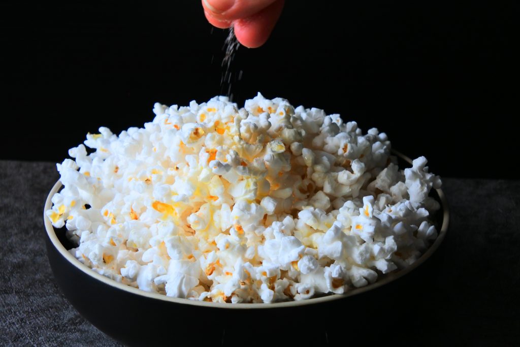 Head on image of a hand sprinkling salt over a bowl of popcorn