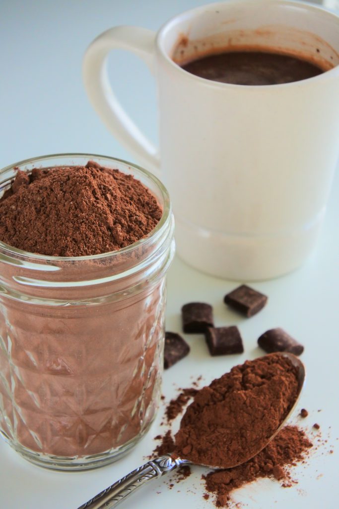 An angled image of a jar of hot cocoa mix with a spoon of hot cocoa and a few chocolate chunks with a white mug of hot cocoa in the background