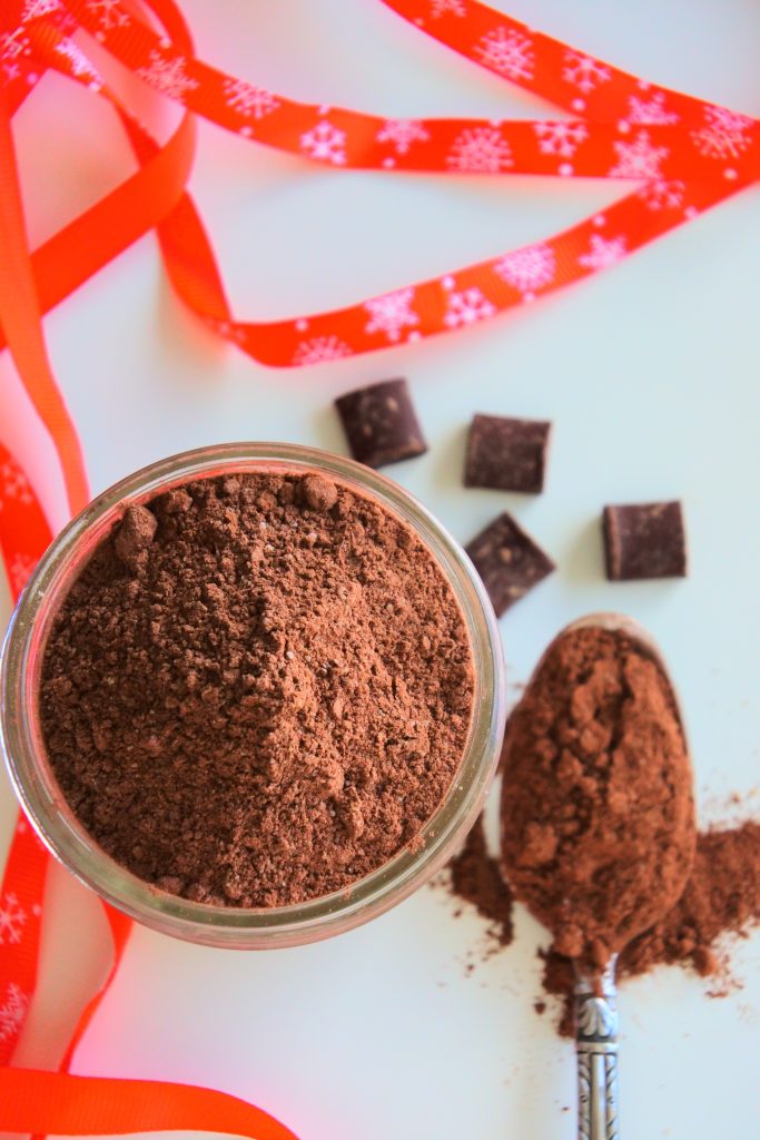 An overhead image of a jar of hot cocoa mix with a spoon of hot cocoa and scattered bits of chocolate surrounded by a red ribbon
