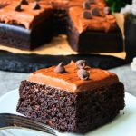 A head on close up image of a square slice of Kitrusy's Healthier Chocolate Cake frosted with chocolate frosting and topped with dark chocolate chips on a plate.