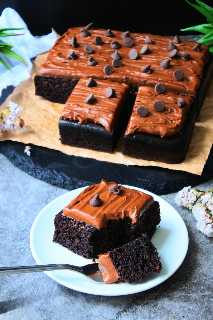 An angled image of Kitrusy's healthier chocolate cake frosted with chocolate frosting and topped with dark chocolate chips with a piece on a plate with a forkful on it