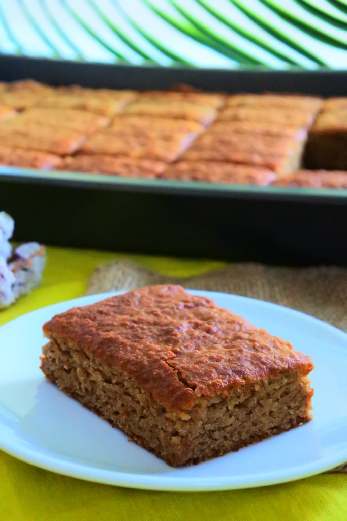A head on image of a piece of warm moist, gluten-free homemade banana bread on a plate with a sheet pan of banana bread in the background.