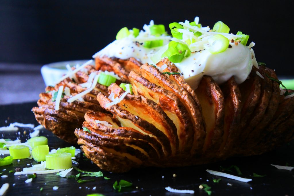 A head on, close up image of a Hasselback potato topped with Greek yogurt, black pepper, spring onions and a sprinkle of Parmesan cheese