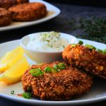 An angled image of two salmon croquettes on a white plate with lemon wedges, chopped spring onions and a small dish of tartar sauce.