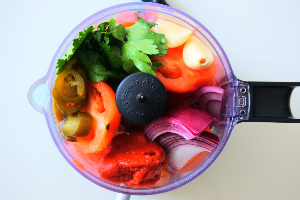 An overhead image of a mini food processor with ingredients needed and added to it for a tomato salsa before being blended together.