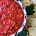 An overhead image of a bowl of tomato salsa in a bowl surrounded by corn chips