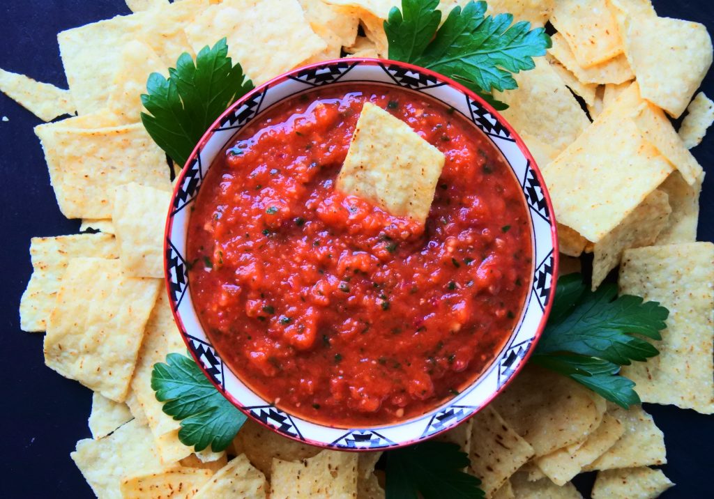 An overhead image of a bowl of fresh tomato salsa surrounded by corn chips on a black slate plate with a single chip dipped into it.