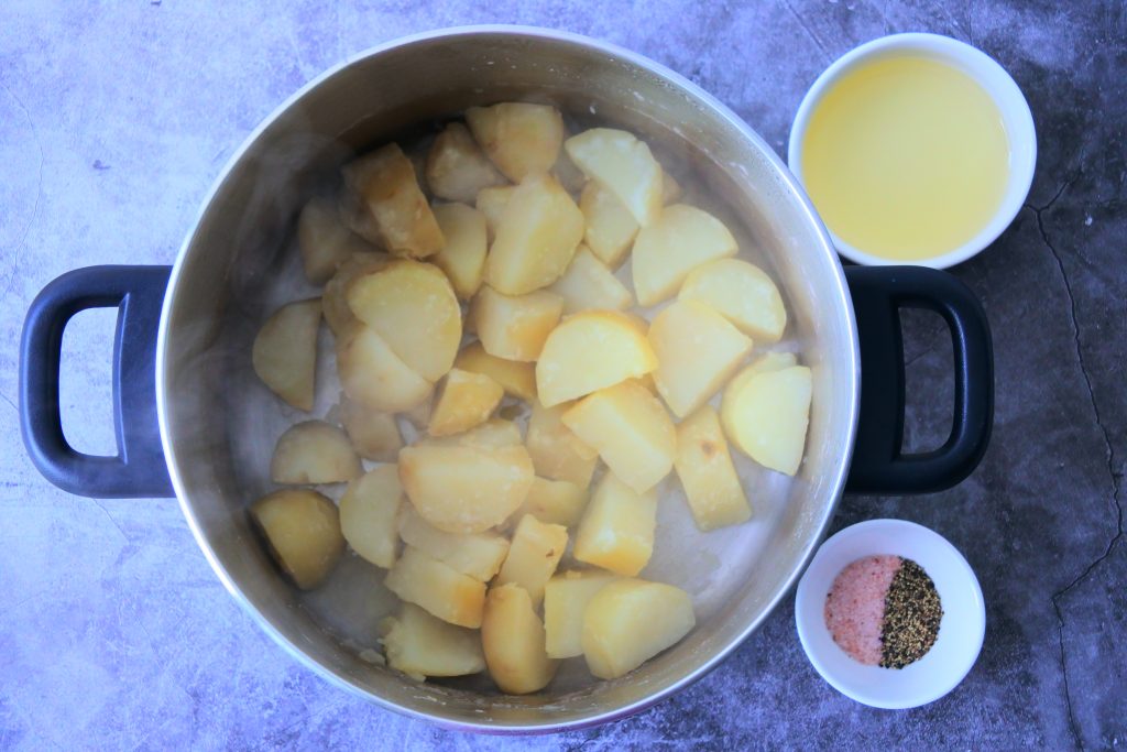 An overhead image of a pot of boiled potato chunks next to a dish of oil and one of salt and pepper