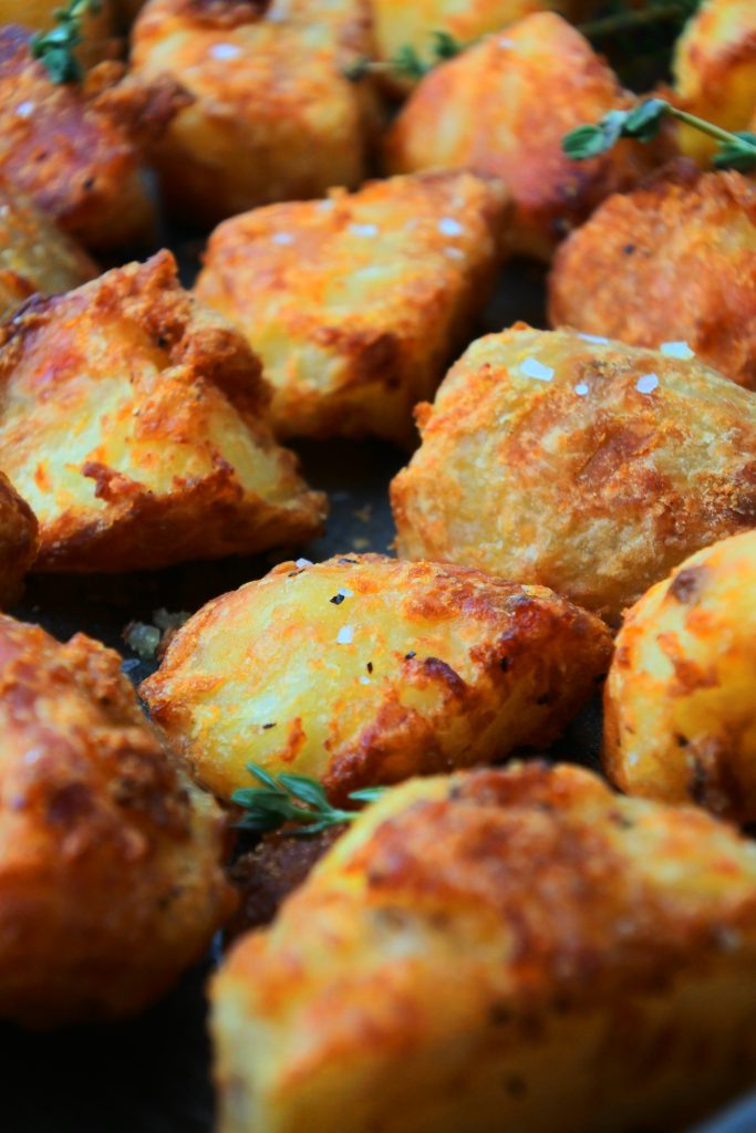 A close up image of crispy roast potatoes on a tray topped with flaky salt and fresh herbs