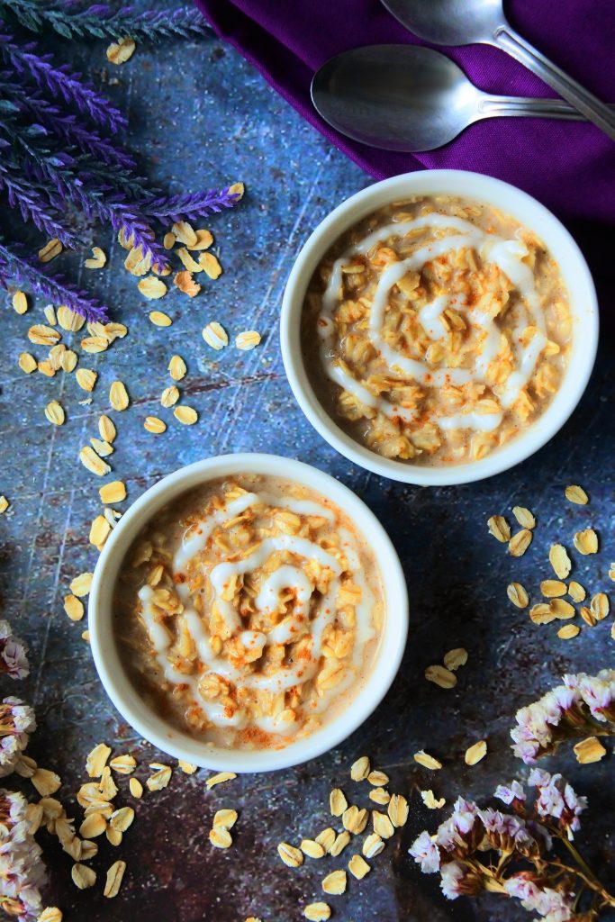 An overhead image showing a two bowls of warm cinnamon roll oatmeal with purple flowers, a purple linen napkin and a silver spoon in the background surrounded by flakes of oats.