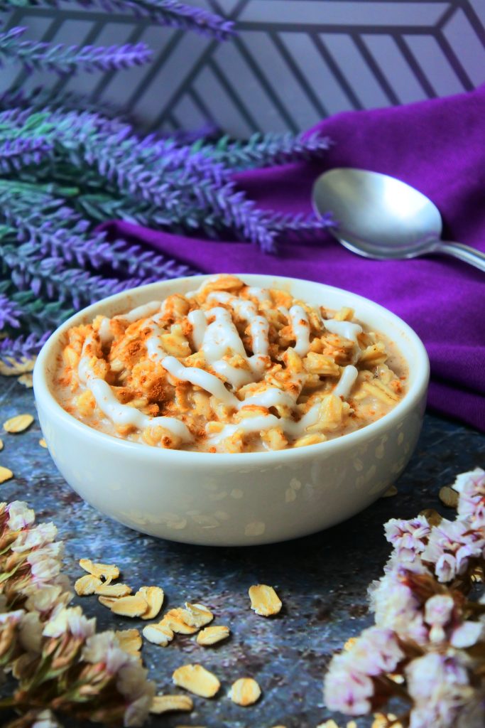 An angled image showing a small bowl of warm cinnamon roll oatmeal with purple flowers, a purple linen napkin and a silver spoon in the background surrounded by flakes of oats.
