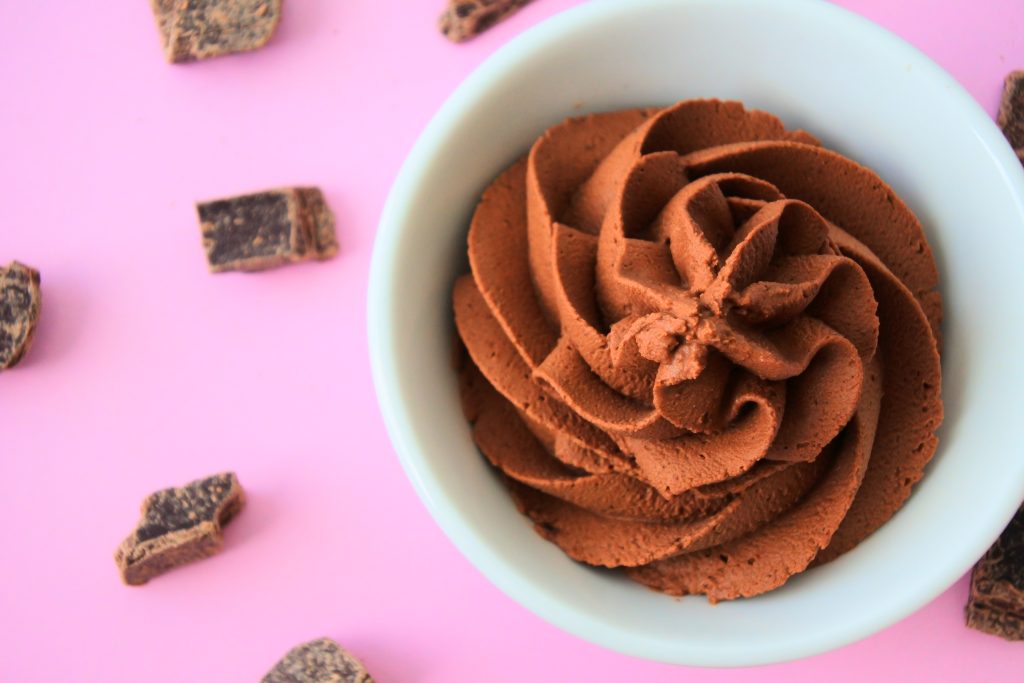 An overhead image of whipped chocolate mousse in a small white dish on a pink background with dark chocolate chunks scattered around