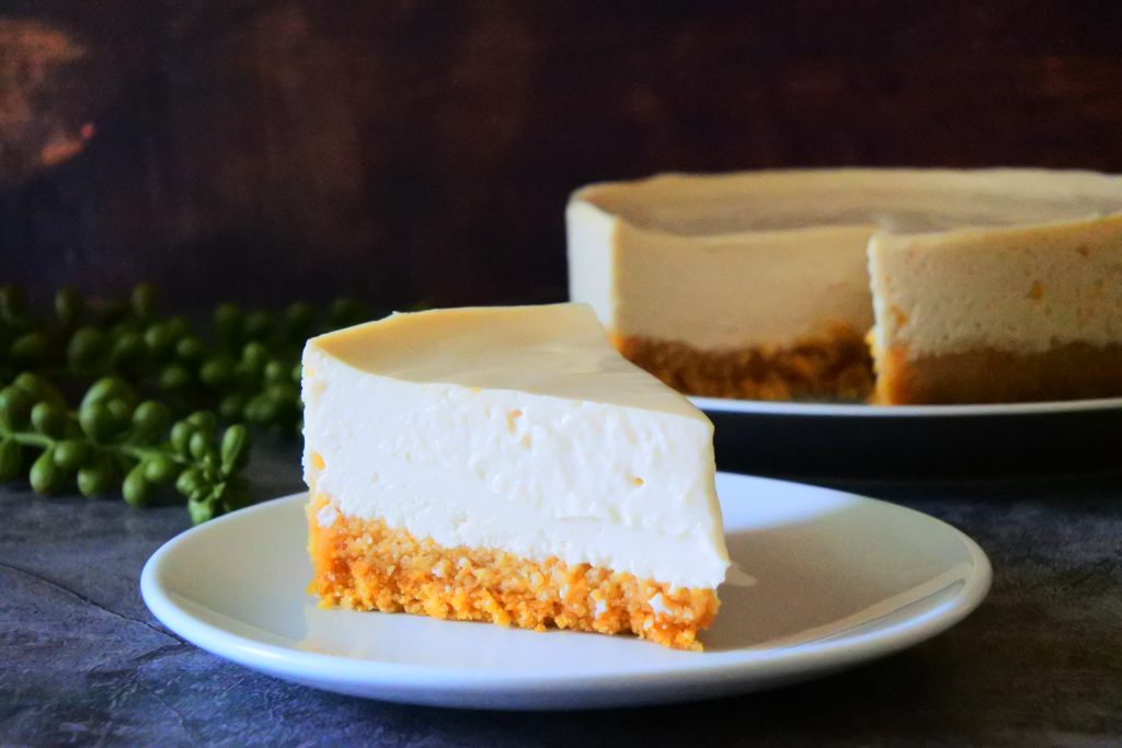 A close up image of a slice of gluten-free Greek yogurt cheesecake on a white plate with the rest of the cheesecake in the background