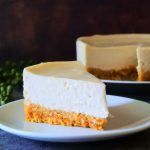 A close up image of a slice of gluten-free Greek yogurt cheesecake on a white plate with the rest of the cheesecake in the background