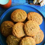An overhead image of a plate of gingerbread cookies surrounded by a cup of milk and fresh spices