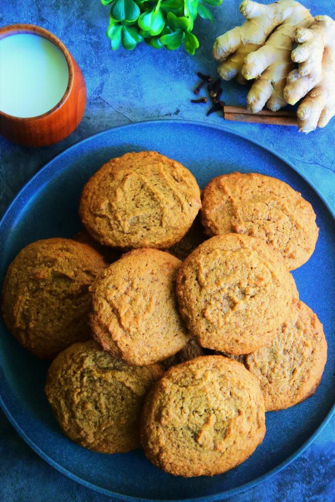 An overhead image of a plate of gingerbread cookies surrounded by a cup of milk and fresh spices