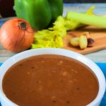 An angled image of a bowl of gravy on a blue linen napkin with a wooden board with onion, celery green pepper, roasted garlic and a carton of broth in the background