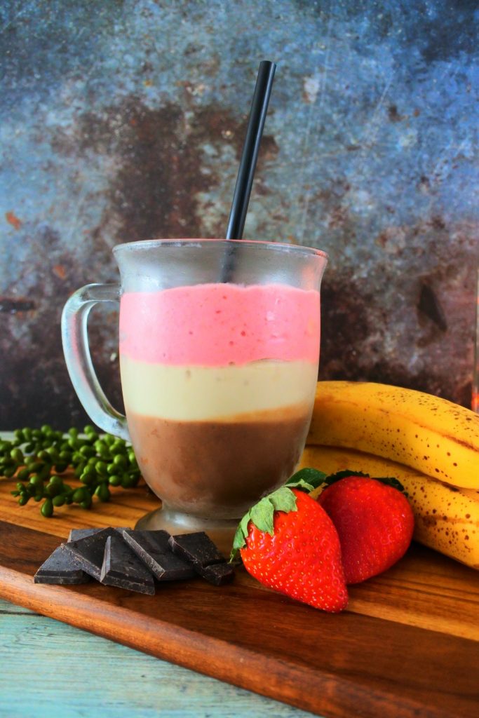 A close up image of a glass mug of Neapolitan Milkshake on a wooden board surrounded by dark chocolate, fresh strawberries and bananas