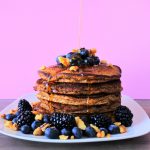 An image of a stack of almond flour pancakes that are topped and surrounded by fruit and nut with a stream of maple syrup being poured onto it.