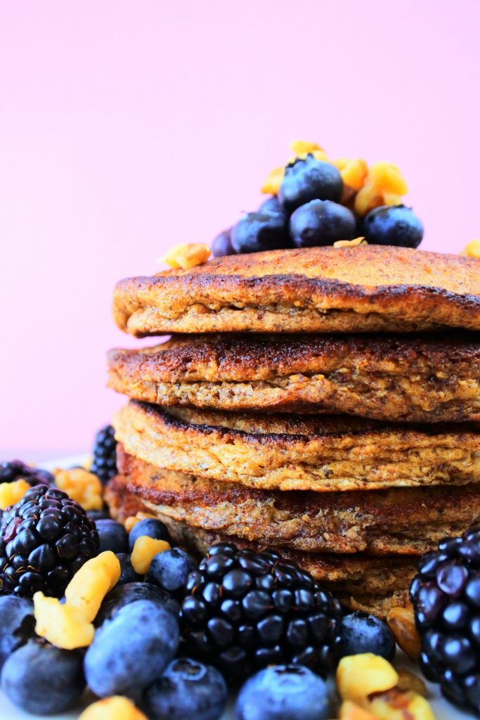 A vertical head-on image of a close up shot of a stack of pancakes topped and surrounded by chopped walnuts, blackberries and blueberries.