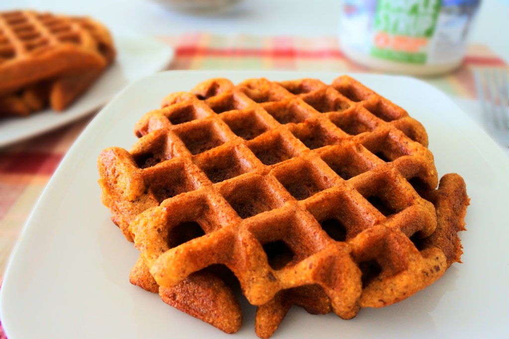 A close up image of almond flour waffles on a plate with a second plate of waffles and a bottle of maple syrup in the background.