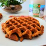 An angled head on image of a plate of waffles with a bottle of maple syrup and a plant in the background